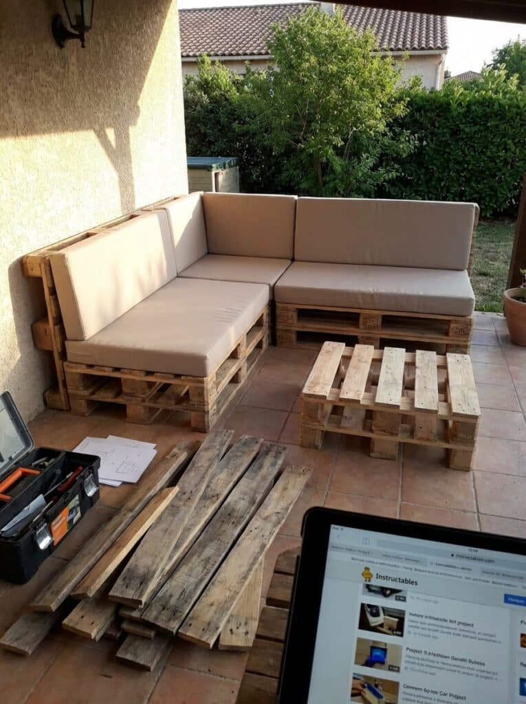 ai-photo-studio-1766941258024-2 DIY outdoor pallet sofa set with beige cushions on a patio, surrounded by wooden planks, tool box, and project instructions. | Sky Rye Design DIY outdoor pallet sofa set with beige cushions on a patio, surrounded by wooden planks, tool box, and project instructions.