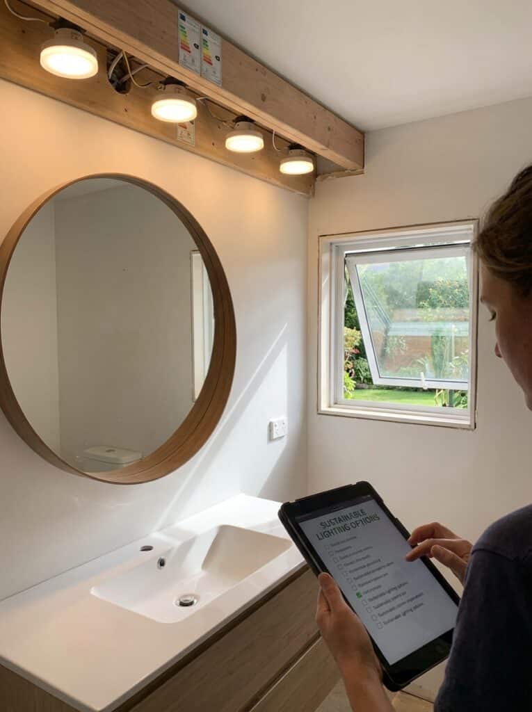 ai-photo-studio-1767012445496-1 Bathroom with eco-friendly lighting, round mirror, window, and person using tablet for sustainable options. | Sky Rye Design Bathroom with eco-friendly lighting, round mirror, window, and person using tablet for sustainable options.