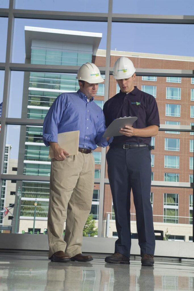 Two construction workers in hard hats review plans indoors against a city building backdrop.