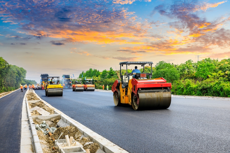 Road construction site with steamrollers paving asphalt under a vibrant sunset sky and surrounded by lush greenery.