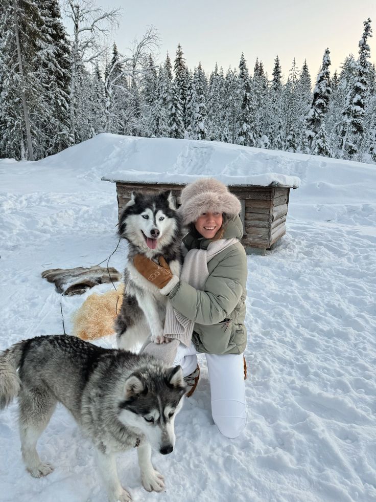 Woman in winter clothing hugging two huskies in a snowy forest setting with a wooden doghouse in the background.