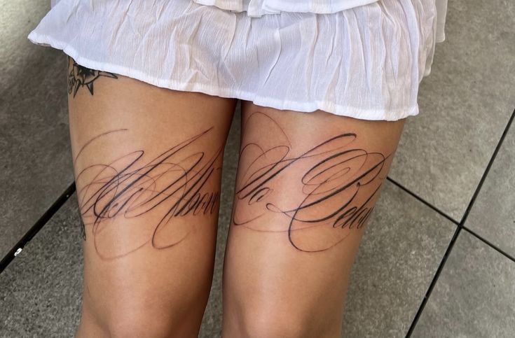 Elegant script tattoos on thighs beneath a white skirt, featuring intricate calligraphy design on a tiled floor background. tattoo font styles