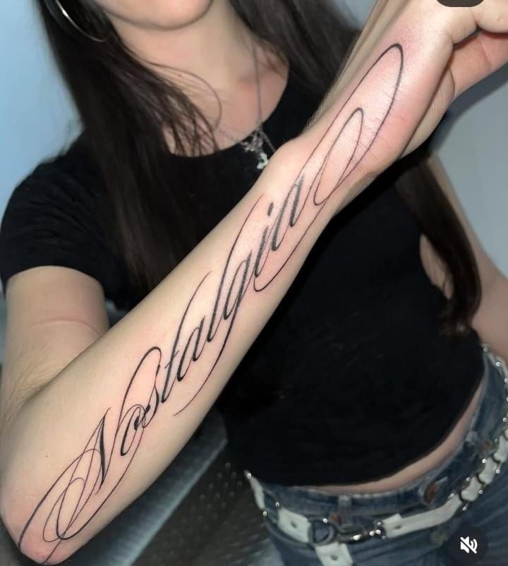 ba06b091-699d-44b1-8248-1fcd1ba4a44a Woman with a Nostalgia tattoo in elegant script on her forearm, wearing a black top and jeans, showcasing artistic body art. | Sky Rye Design Woman with a Nostalgia tattoo in elegant script on her forearm, wearing a black top and jeans, showcasing artistic body art.