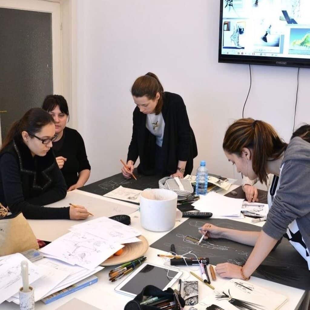 bb3debc1f14866f2a065eee4ee31e01e Group of focused students drawing and collaborating at a table filled with sketches, pencils, and a monitor displaying reference images. | Sky Rye Design Group of focused students drawing and collaborating at a table filled with sketches, pencils, and a monitor displaying reference images.