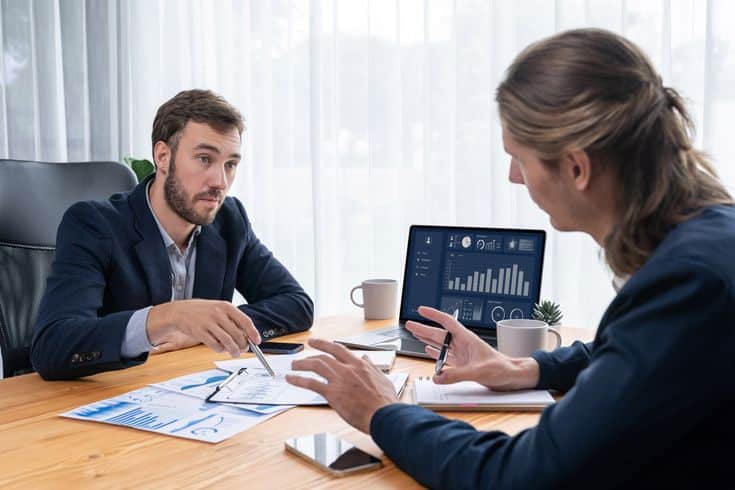 Two business professionals discussing data charts and graphs on papers and a laptop in an office setting with natural light.