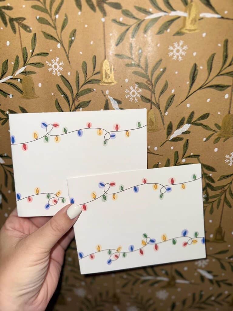 c9c21d40509050f49bc2a87b920c459e Hand holding greeting cards with colorful Christmas lights, set against festive wrapping paper with snowflakes and winter foliage. | Sky Rye Design Hand holding greeting cards with colorful Christmas lights, set against festive wrapping paper with snowflakes and winter foliage.