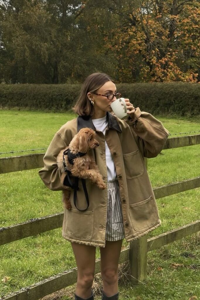 Screenshot Woman in jacket holding a small dog and sipping coffee outdoors in autumn, surrounded by lush greenery and a rustic wooden fence. | Sky Rye Design Woman in jacket holding a small dog and sipping coffee outdoors in autumn, surrounded by lush greenery and a rustic wooden fence.