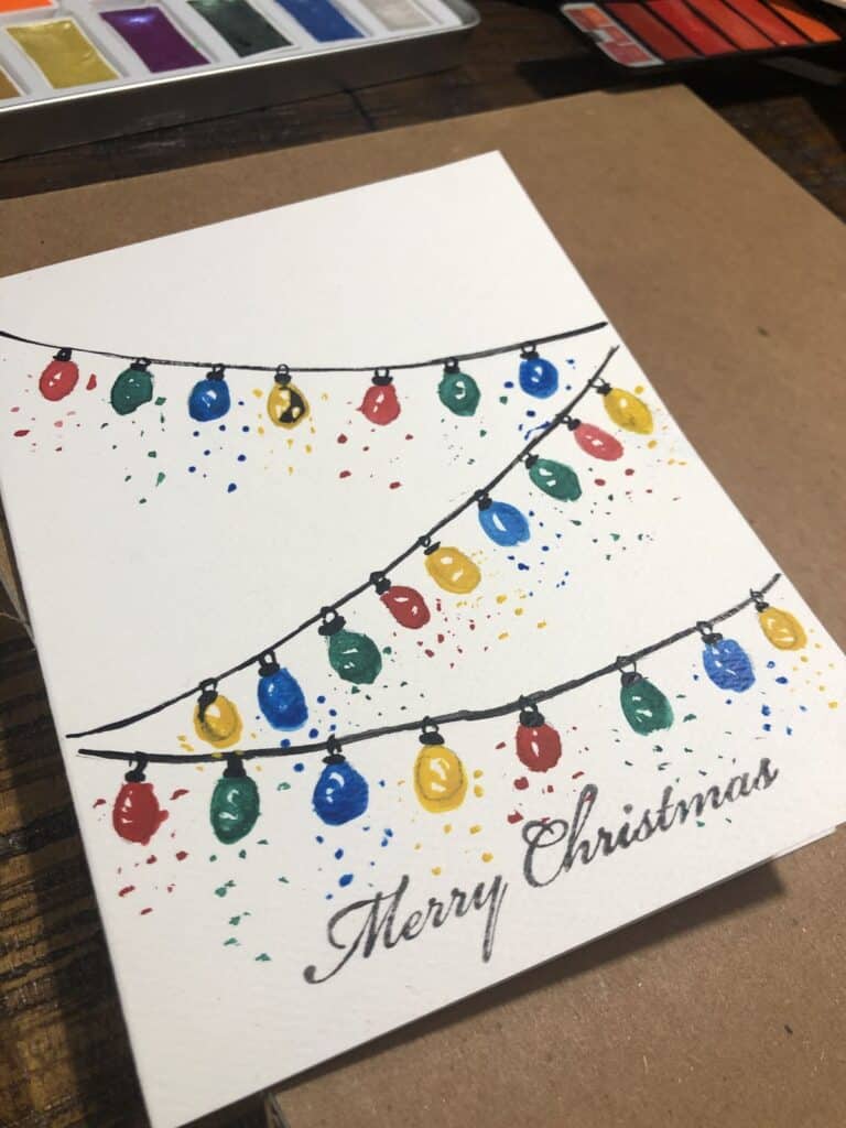 cf60dea616243a5e55aac7bf61d59d39 Watercolor Christmas card with colorful string lights and Merry Christmas message, on a rustic wooden table with paint set. | Sky Rye Design Watercolor Christmas card with colorful string lights and Merry Christmas message, on a rustic wooden table with paint set.