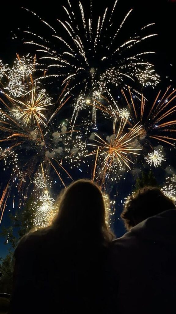 A couple watches vibrant fireworks fill the night sky, celebrating a festive occasion outdoors.