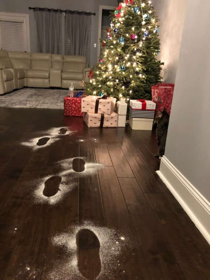 Footprints in snow dust lead to a Christmas tree with glowing lights and wrapped gifts, creating a festive holiday atmosphere.