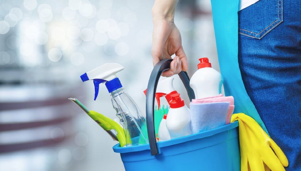 Person carrying a blue cleaning supply caddy with spray bottles, sponges, and rubber gloves, ready for household chores or cleaning tasks.