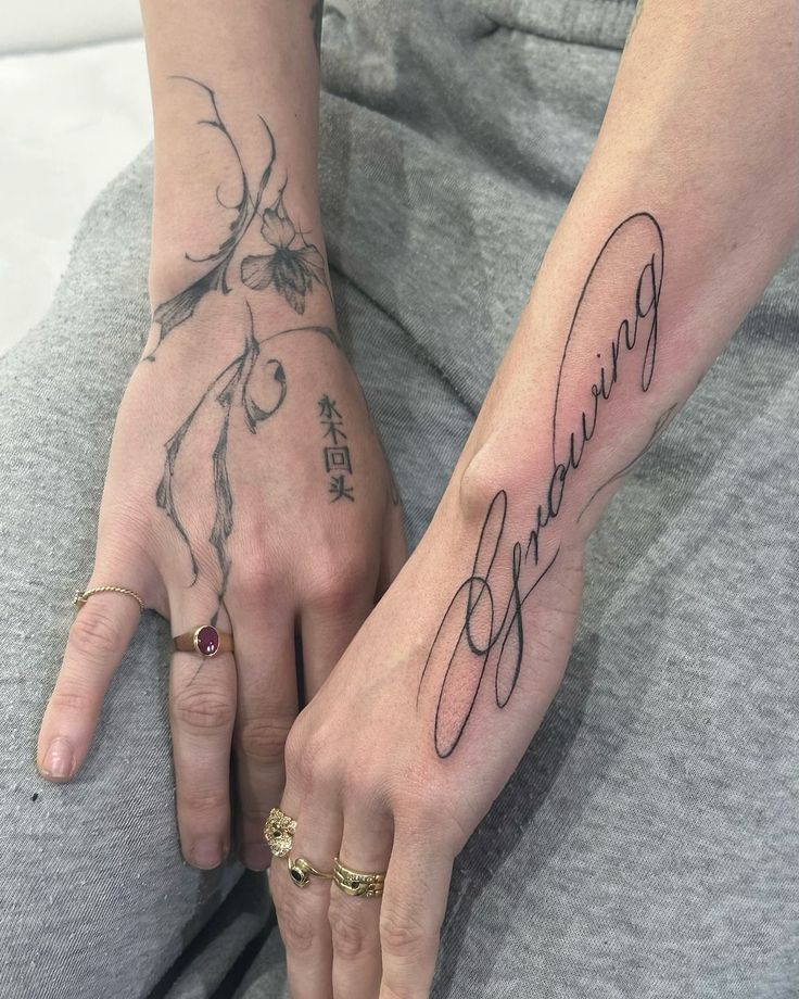 cursive lettering Close-up of hands with artistic tattoos and rings, one showcasing floral designs and the other featuring the word growing in elegant script. | Sky Rye Design Close-up of hands with artistic tattoos and rings, one showcasing floral designs and the other featuring the word growing in elegant script.