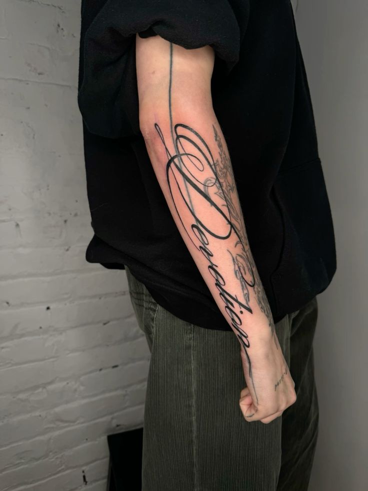 Forearm tattoo with intricate black script reading Devotion on a person's arm, set against a brick wall background. tattoo fonts for names