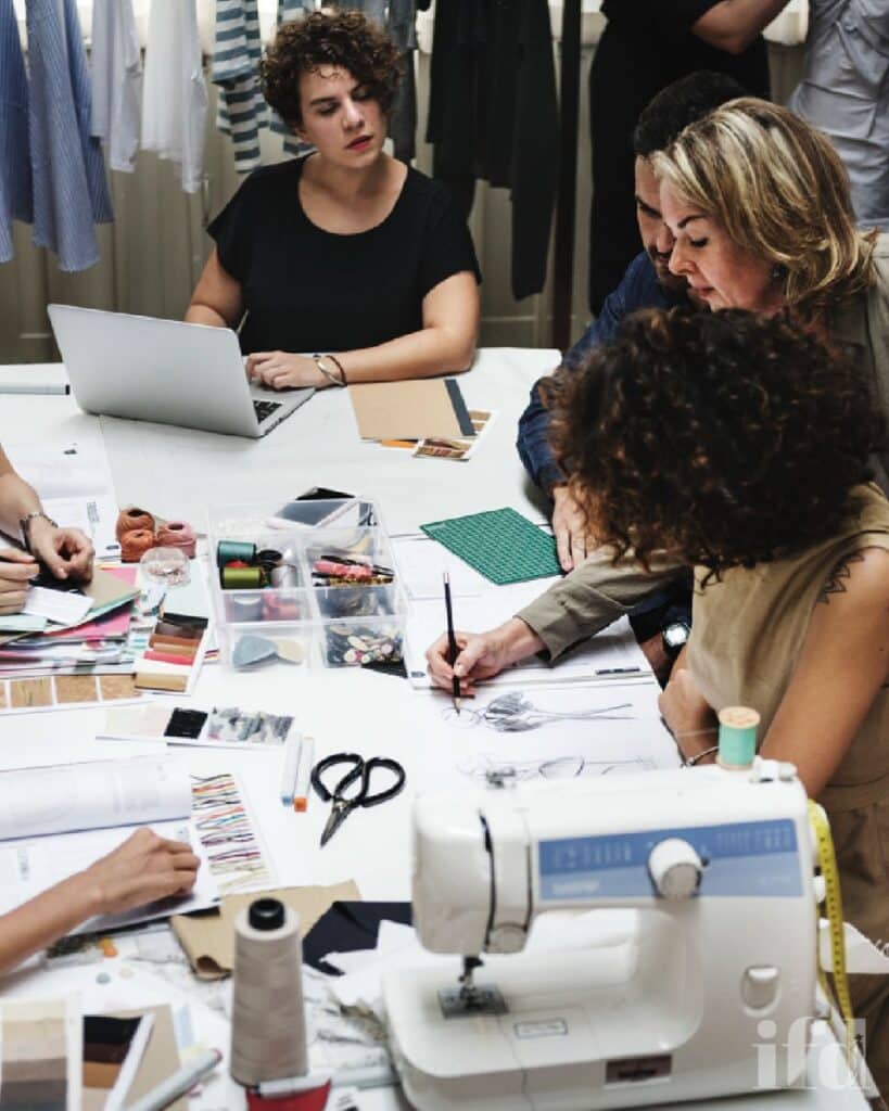 Fashion design team collaborating at a table with sewing machine, fabric samples, and sketches. Creative teamwork in a design studio.