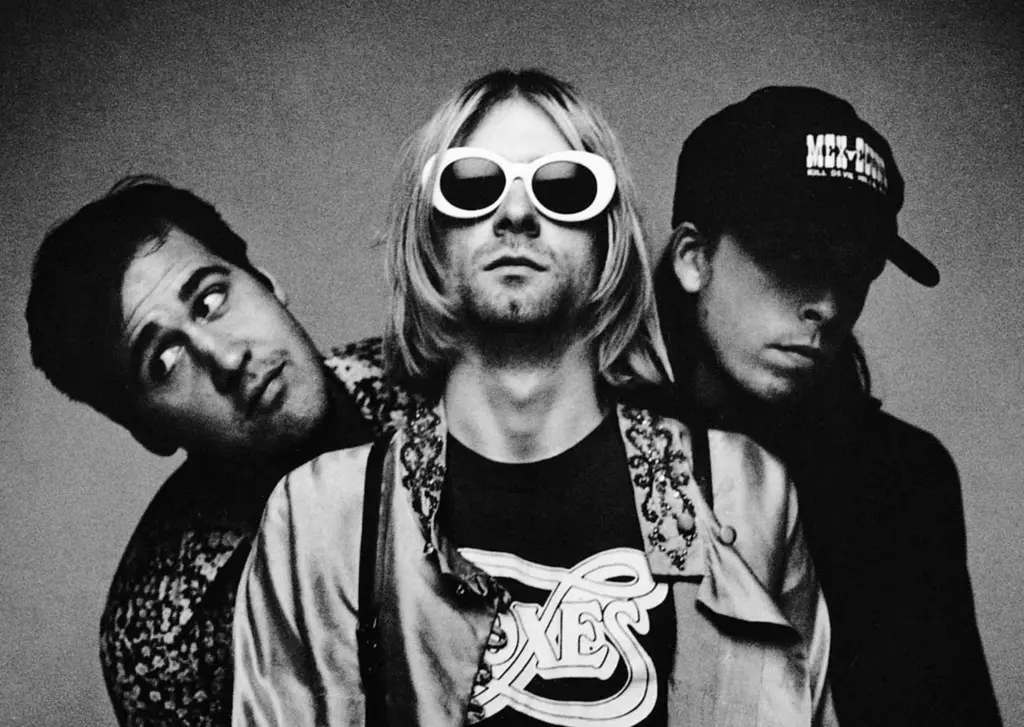 Black-and-white portrait of three 1990s grunge band members; center singer wearing white oval sunglasses