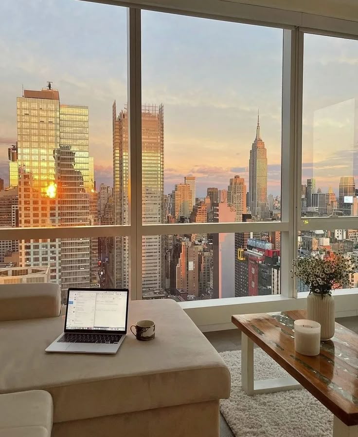 Modern living room with laptop and coffee overlooking New York City skyline at sunset through large windows. Cozy workspace ambiance.