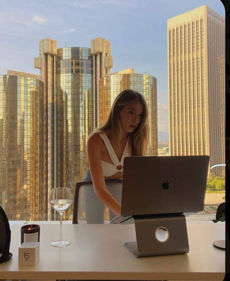 Woman working on a laptop in a modern office with cityscape view, featuring tall glass buildings and a glass of wine on the desk.