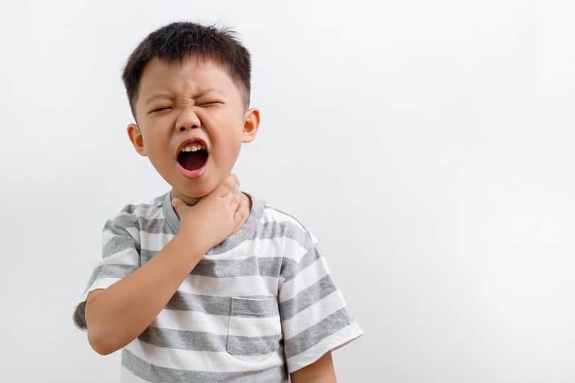 Child holding neck, expressing discomfort, white background, wearing gray-striped shirt, indicating sore throat or difficulty swallowing.