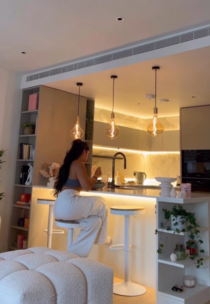 ee0a9ad4-dd3e-4c8b-ada0-1bcbe49a4718 Woman sitting at a modern kitchen counter with ambient lighting and stylish decor, enjoying a meal under pendant lights. | Sky Rye Design Woman sitting at a modern kitchen counter with ambient lighting and stylish decor, enjoying a meal under pendant lights.
