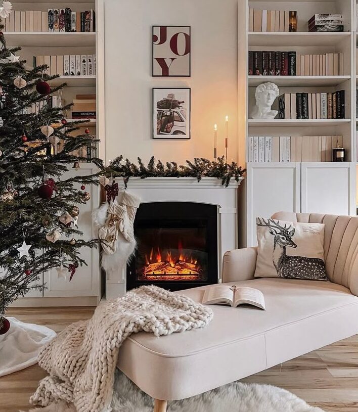 ef100558-0d80-46ac-9e23-dc58c309405a Cozy holiday living room with Christmas tree, decorated fireplace, and bookshelves. Soft sofa with pillows and a blanket nearby. | Sky Rye Design Cozy holiday living room with Christmas tree, decorated fireplace, and bookshelves. Soft sofa with pillows and a blanket nearby.