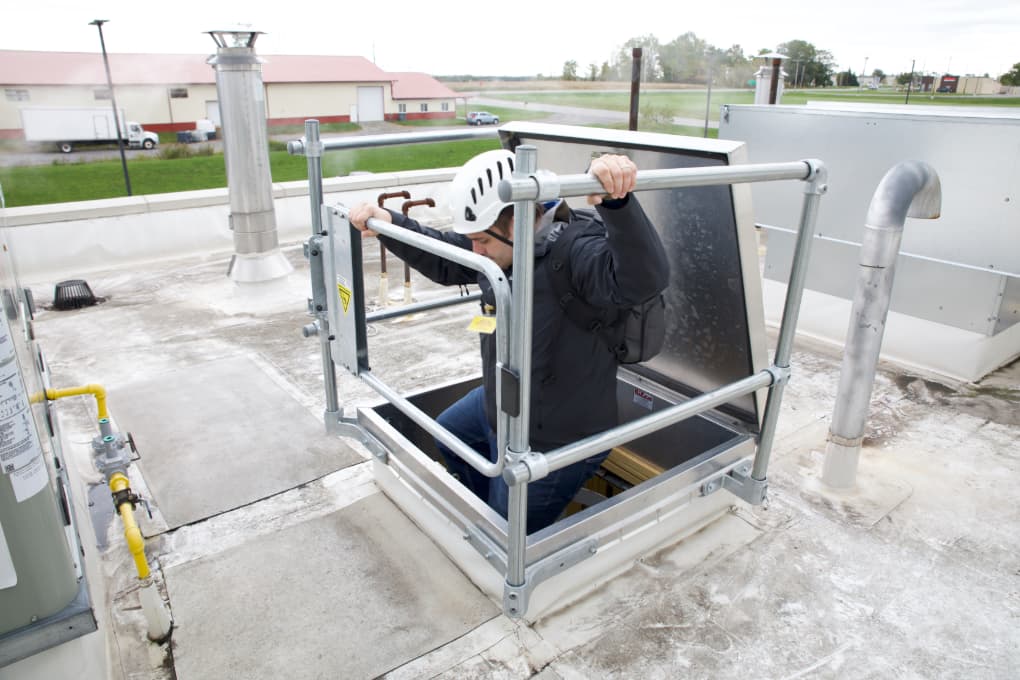 Person wearing helmet safely exiting roof hatch with protective railing on industrial building rooftop.