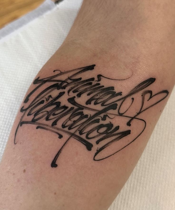 Intricate black tattoo on forearm with artistic script lettering and decorative elements, displayed on a light skin background.