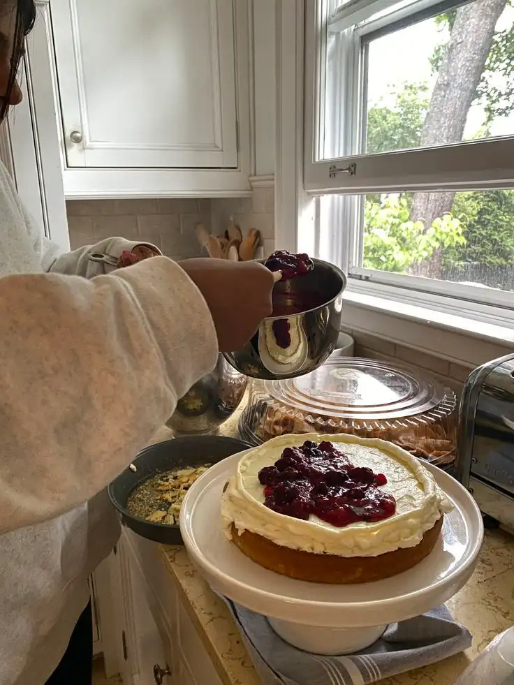 Person adding berry topping to a cheesecake on a kitchen counter near a window, creating a delicious homemade dessert.