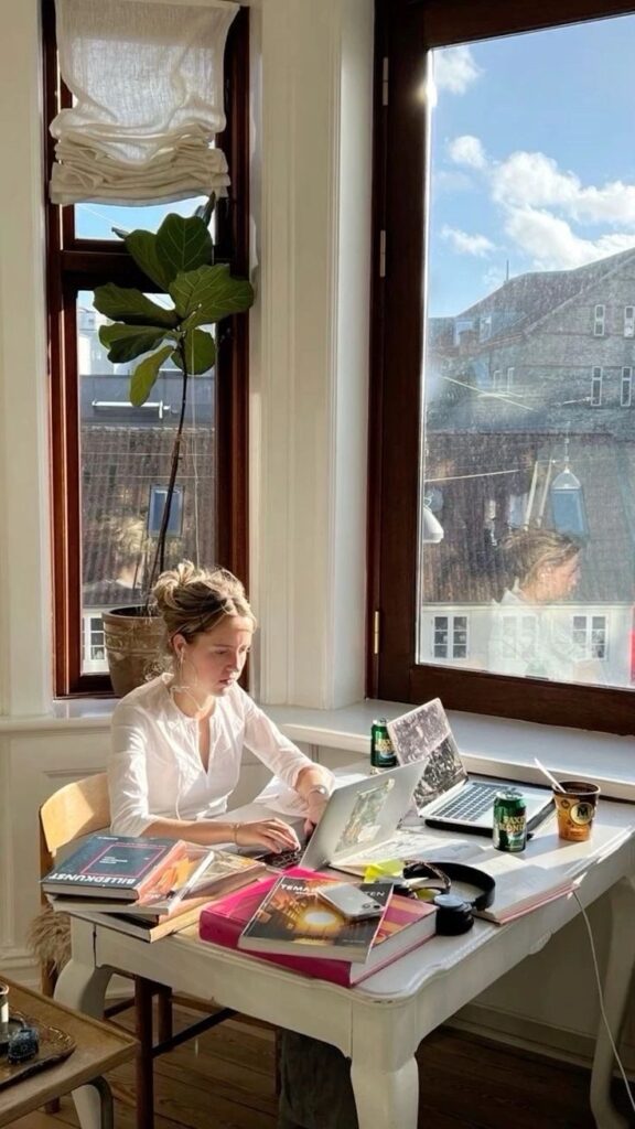 Woman working at a sunlit desk with books, laptop, and drinks. Cozy home office with natural light and outdoor view.