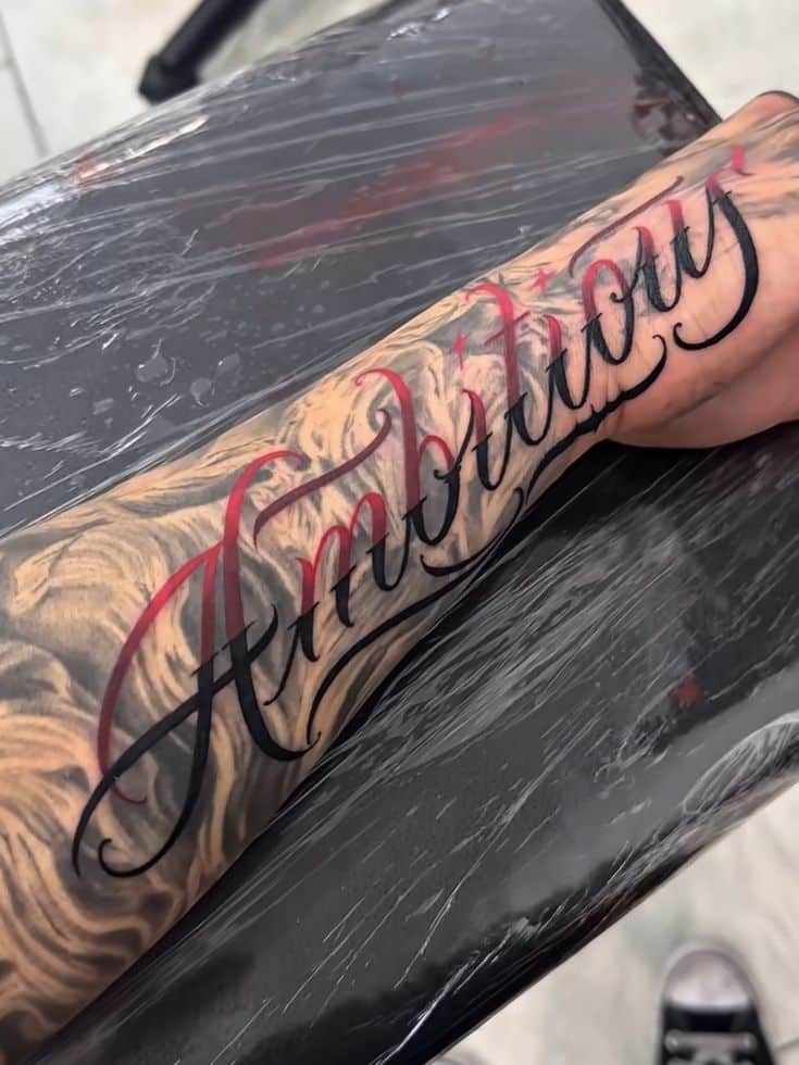f36697bc-120f-45e1-bd3d-7f881cad89b2 Tattooed arm with the word Ambitious in elegant script, surrounded by intricate shading, resting on a surface covered in plastic wrap. | Sky Rye Design Tattooed arm with the word Ambitious in elegant script, surrounded by intricate shading, resting on a surface covered in plastic wrap.