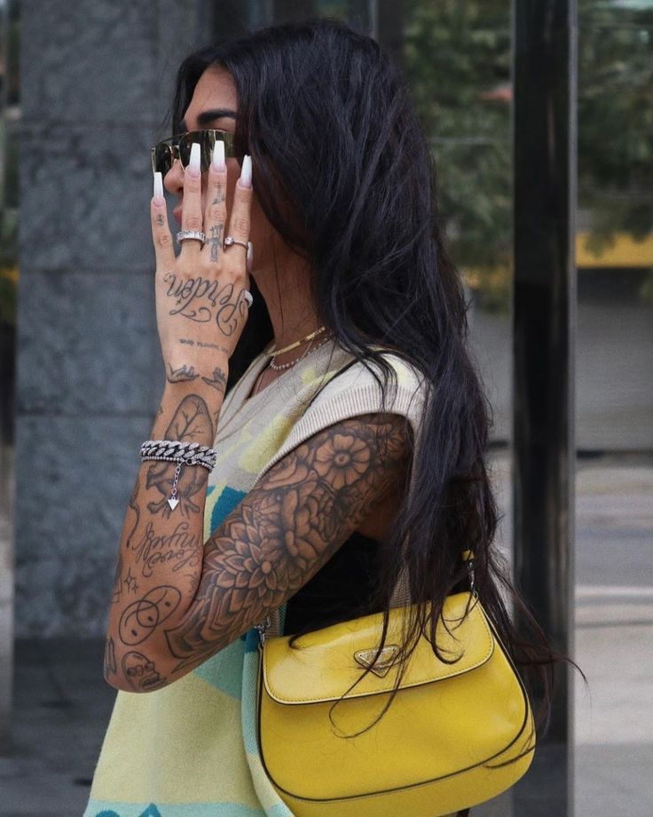 Stylish woman with tattoos and sunglasses holds a yellow handbag, showcasing trendy accessories and fashion-forward style.