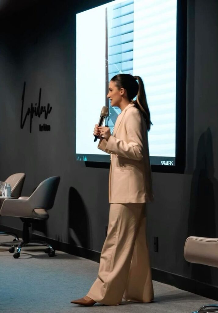 Professional woman in beige suit gives a presentation with a microphone, standing in front of a screen in a conference room.