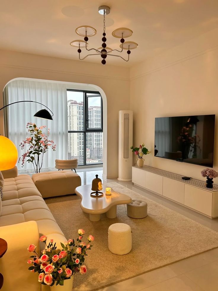 fe9489fa-db66-40d1-a652-ae38cdedbff1 Modern living room with cream furniture, large TV, lush plants, and floor-to-ceiling windows showcasing urban skyline. Cozy and elegant design. | Sky Rye Design Modern living room with cream furniture, large TV, lush plants, and floor-to-ceiling windows showcasing urban skyline. Cozy and elegant design.