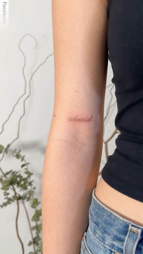 Minimalist celestial tattoo on inner arm in cursive script, woman wearing black top and jeans, with plant in the background.