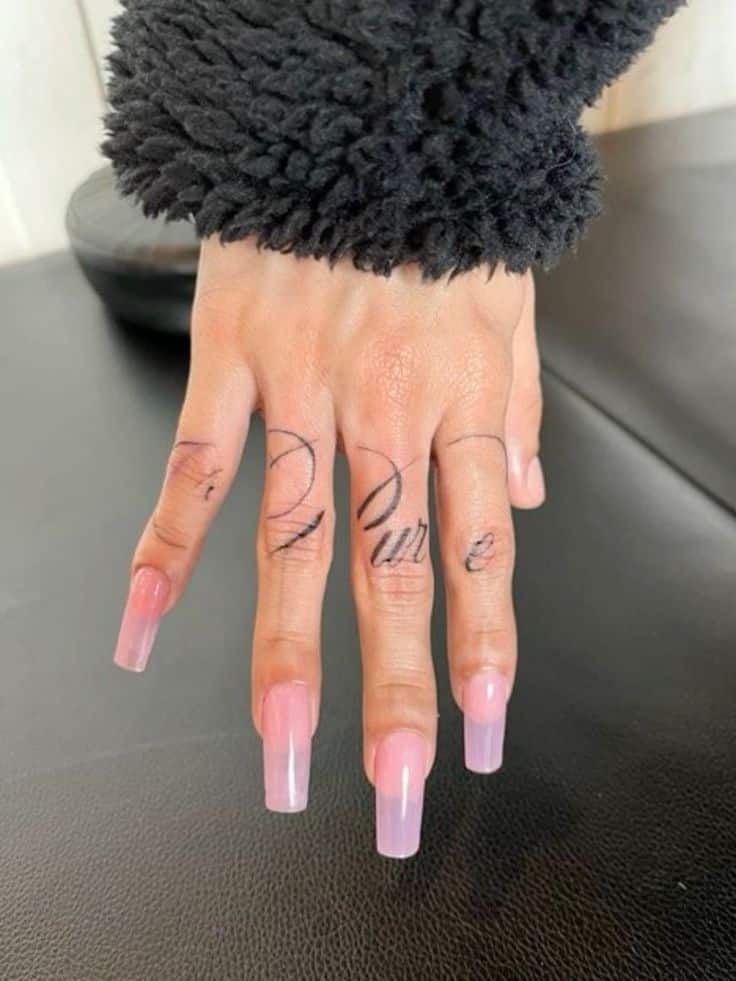 font cursive tattoo _ script tattoo _pure Close-up of hand with long pink acrylic nails and finger tattoos, wearing a black fuzzy sweater on a dark surface. | Sky Rye Design Close-up of hand with long pink acrylic nails and finger tattoos, wearing a black fuzzy sweater on a dark surface.