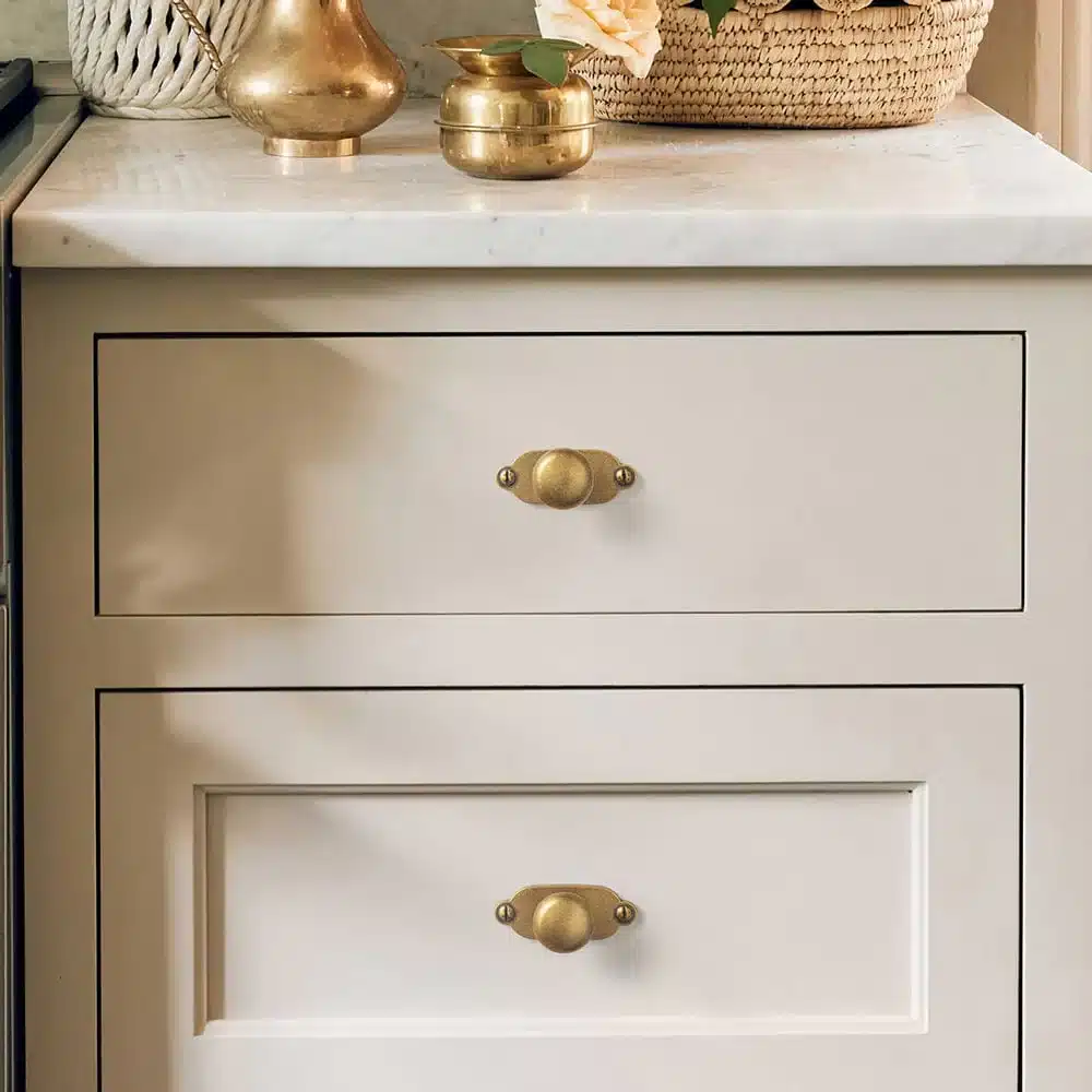 White cabinet with brass handles, topped with decorative brass vases and a woven basket. Stylish and elegant kitchen decor.