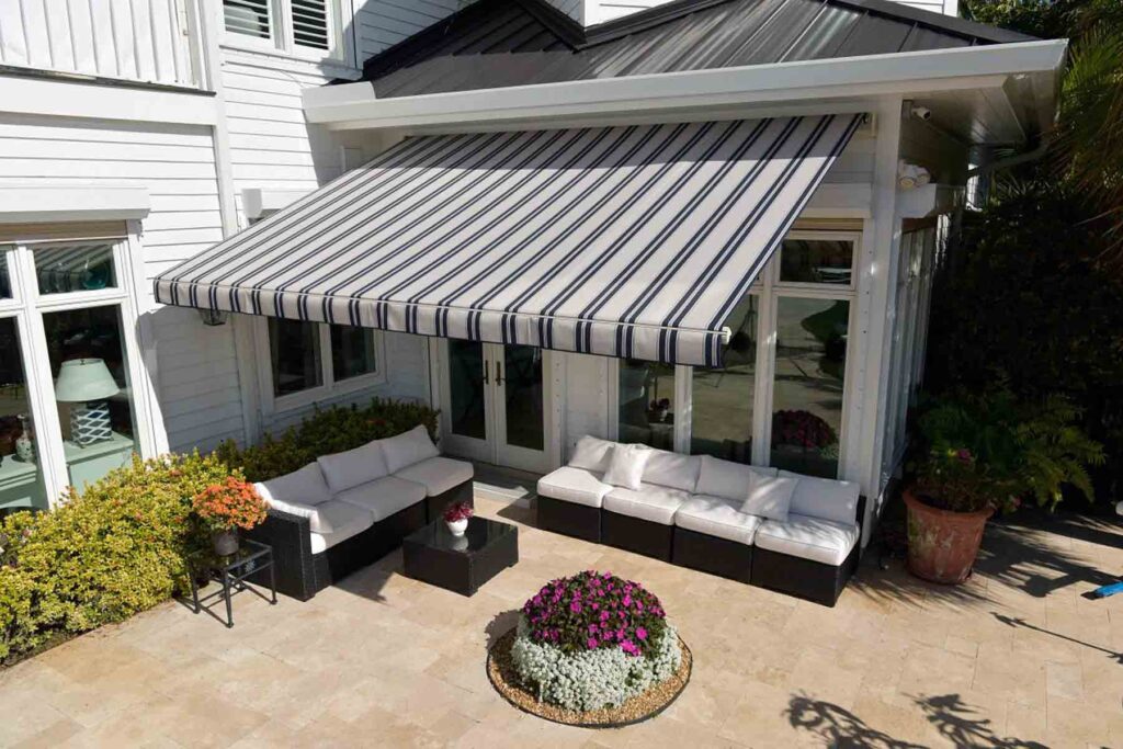 homeguide-sunsetter-platinum-awning Striped awning over a bright patio with white sofas, tables, and vibrant flowers, attached to a modern house exterior. | Sky Rye Design Striped awning over a bright patio with white sofas, tables, and vibrant flowers, attached to a modern house exterior.