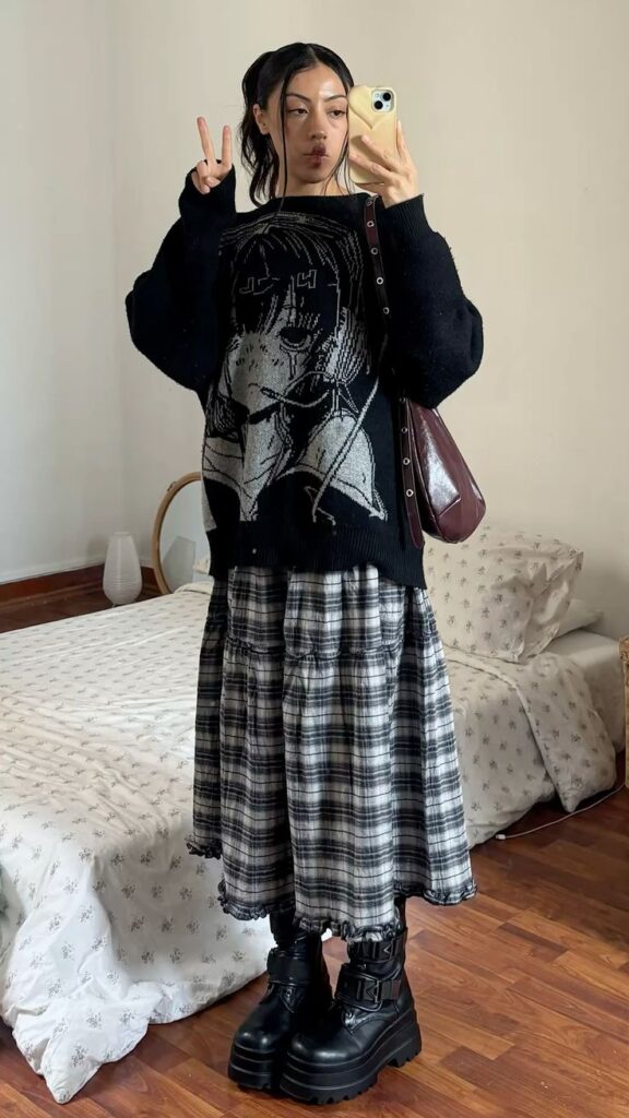 Young woman in stylish anime sweater and plaid skirt poses for mirror selfie, flashing a peace sign. Edgy fashion vibe in cozy bedroom.