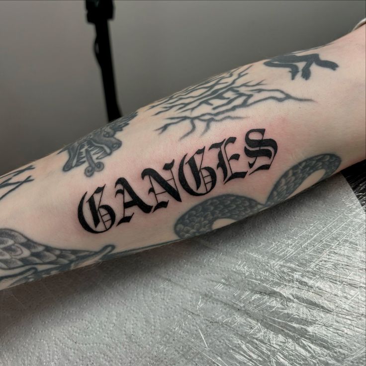 Tattooed forearm featuring the word GANGES in bold, gothic font surrounded by various black ink designs on pale skin.