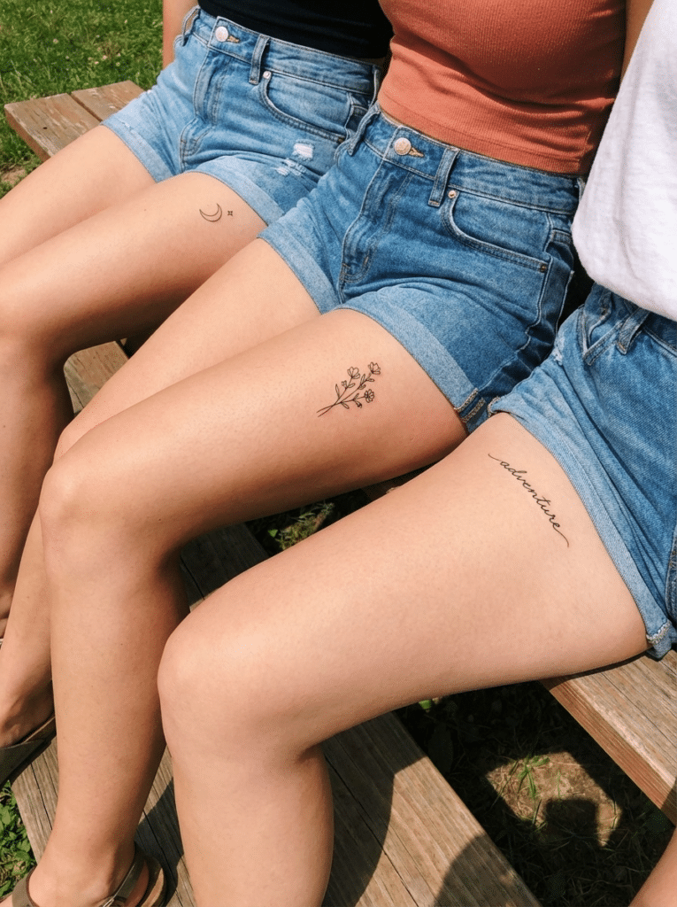 image Three women sitting with thigh tattoos in denim shorts: moon, flowers, and 'adventure'. Shot outdoors on a bench. | Sky Rye Design Three women sitting with thigh tattoos in denim shorts: moon, flowers, and 'adventure'. Shot outdoors on a bench.