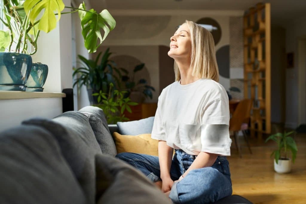 Woman enjoying morning sunlight on couch at home, surrounded by plants, engaging in a mindful moment.