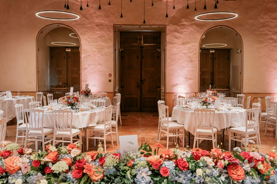Elegant wedding reception with round tables, white chairs, and vibrant floral centerpieces in a warmly lit venue.
