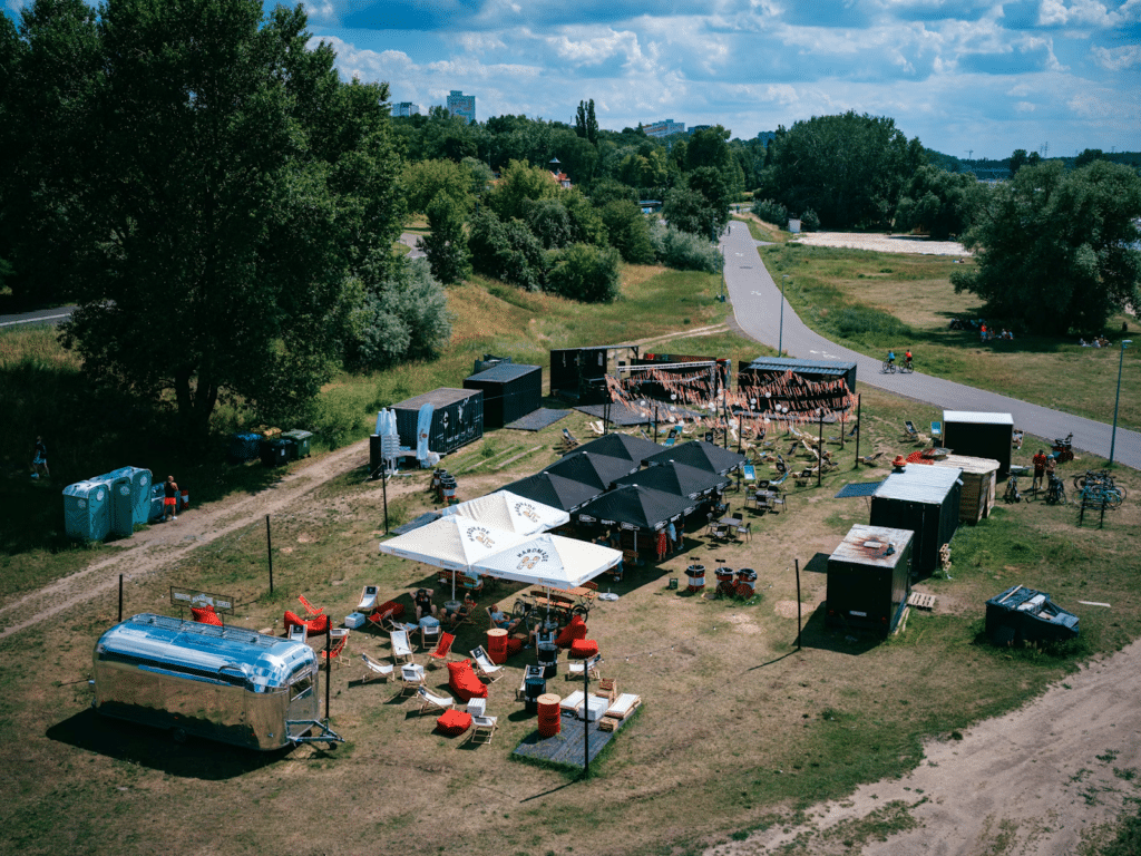 Outdoor festival area with tent, seating, and a trailer on a sunny day, surrounded by greenery and a bike path in the distance.