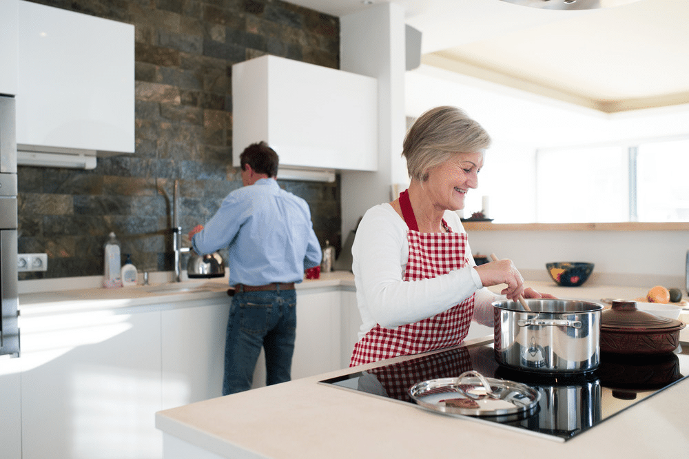 Senior woman cooking in modern kitchen, wearing red checkered apron, while a man rinses dishes in the background near the sink.
