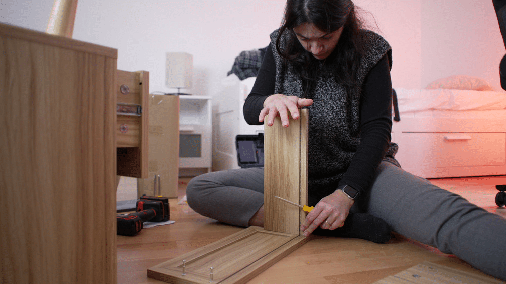 image Woman assembling wooden furniture with a screwdriver on the floor, DIY home project in a cozy bedroom setting. | Sky Rye Design Woman assembling wooden furniture with a screwdriver on the floor, DIY home project in a cozy bedroom setting.