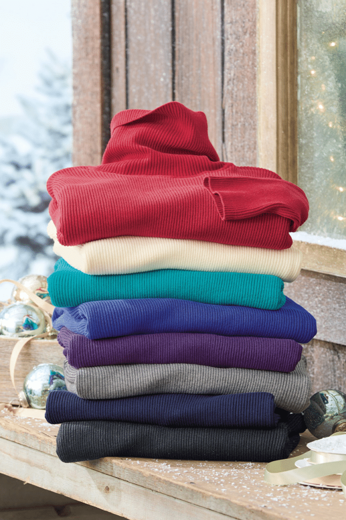 image Stack of vibrant ribbed sweaters in various colors, perfect for winter, displayed on a snowy wooden surface near festive ornaments. | Sky Rye Design Stack of vibrant ribbed sweaters in various colors, perfect for winter, displayed on a snowy wooden surface near festive ornaments.