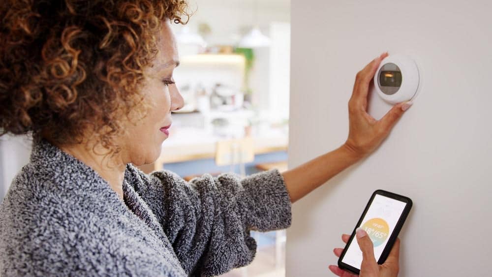 image Woman adjusting smart thermostat with smartphone app for efficient home heating control. | Sky Rye Design Woman adjusting smart thermostat with smartphone app for efficient home heating control.