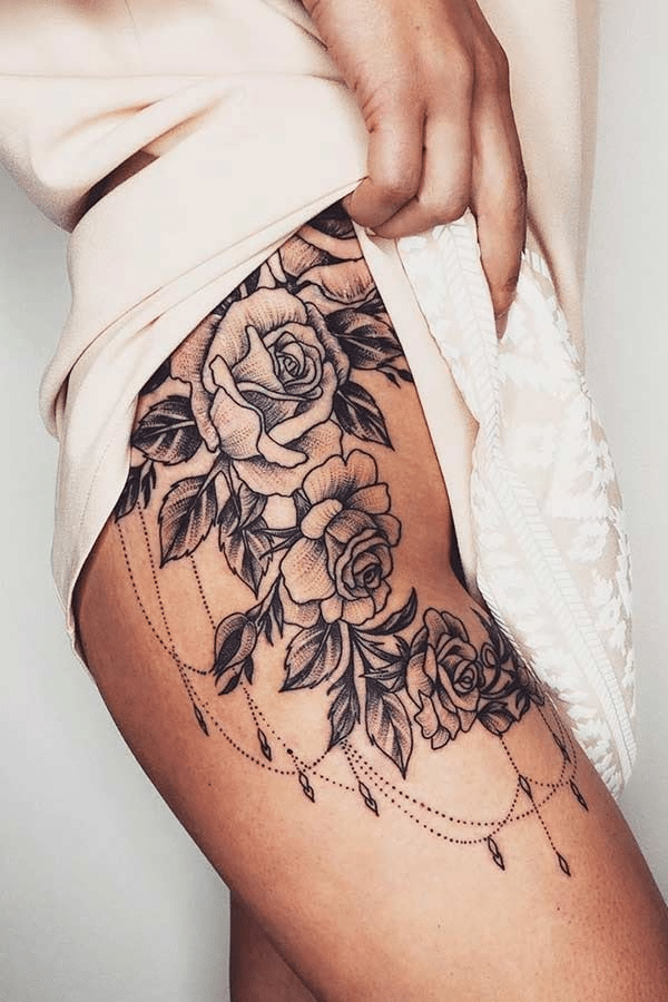 image Floral thigh tattoo featuring detailed roses and decorative linework on a woman's leg, partially visible under cream-colored fabric. | Sky Rye Design Floral thigh tattoo featuring detailed roses and decorative linework on a woman's leg, partially visible under cream-colored fabric.