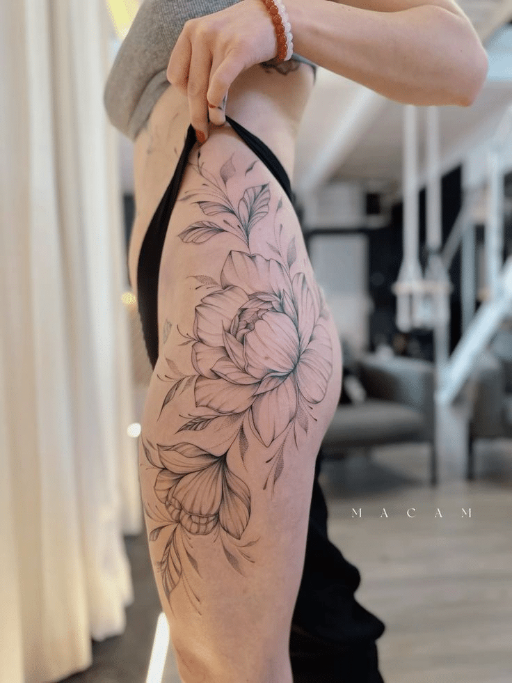 image Detailed black and gray floral thigh tattoo on a person standing indoors, showcasing intricate petal and leaf designs. | Sky Rye Design Detailed black and gray floral thigh tattoo on a person standing indoors, showcasing intricate petal and leaf designs.