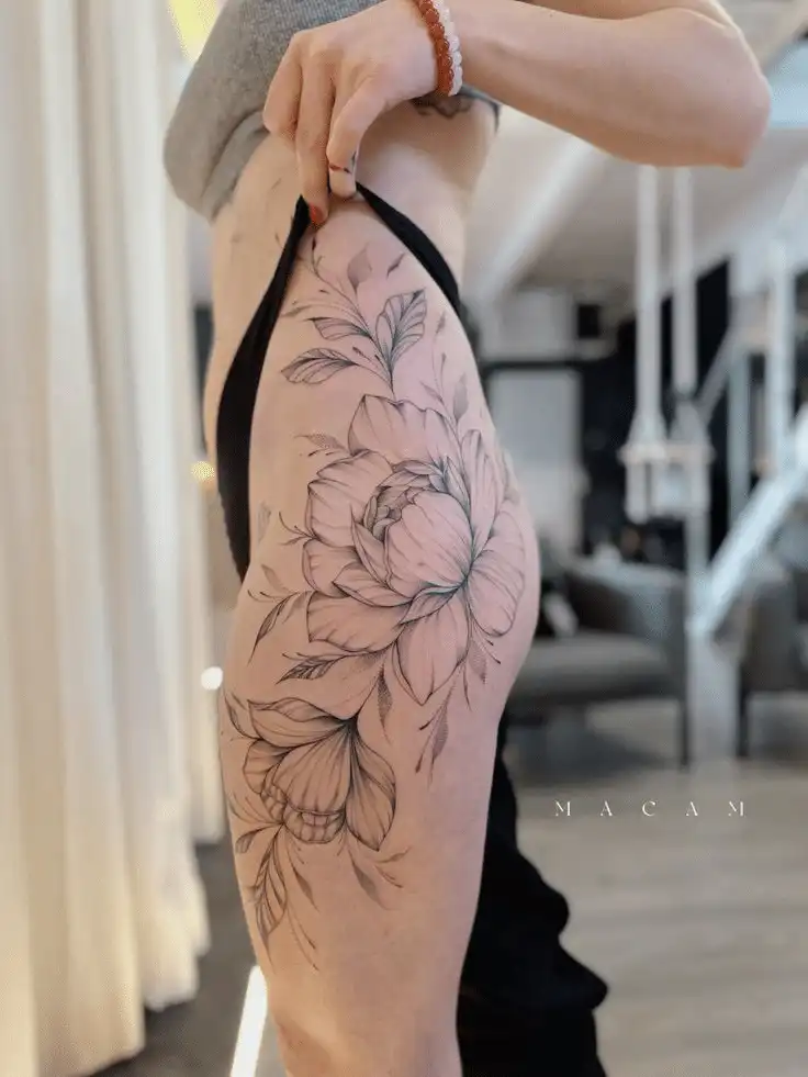 Detailed black and gray floral thigh tattoo on a person standing indoors, showcasing intricate petal and leaf designs.