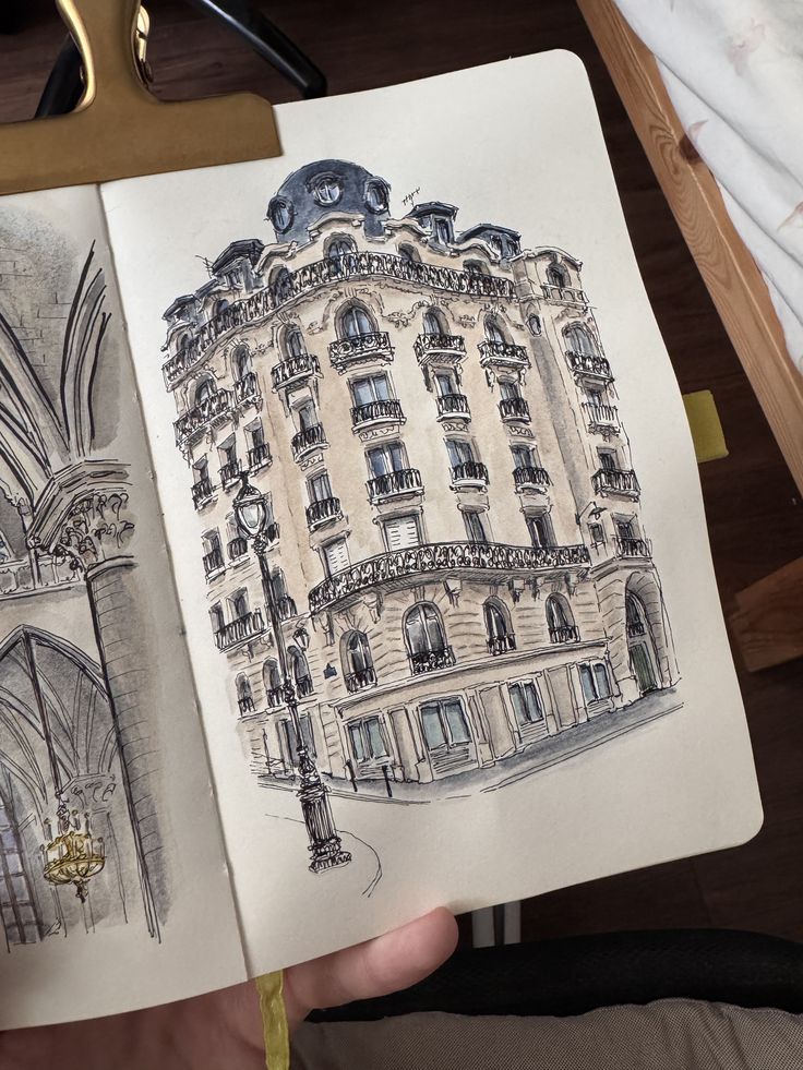 itsninel Architectural sketch of an elegant corner building with ornate balconies, drawn in a sketchbook held open, showcasing artistic detail. | Sky Rye Design Architectural sketch of an elegant corner building with ornate balconies, drawn in a sketchbook held open, showcasing artistic detail.